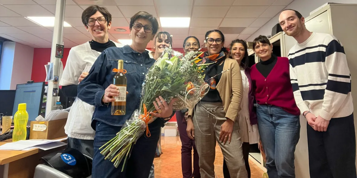 Groupe de personnes souriantes dans un bureau, une femme au centre tenant un bouquet de fleurs et une bouteille